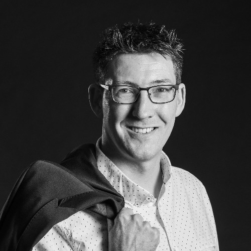 Portrait en noir et blanc d'un homme souriant, à lunettes, une veste sur l'épaule.